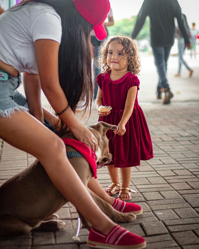A young girl smiles at a friendly dog sitting beside a woman in a busy city area.