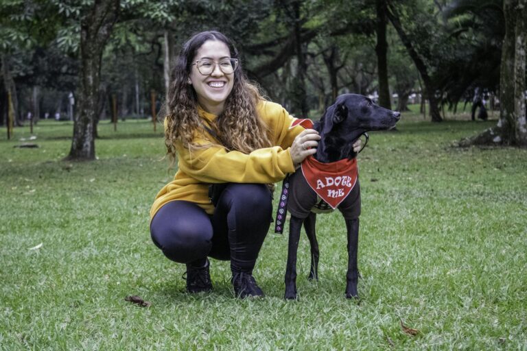 Smiling woman with adoptable black dog in a lush green park, promoting pet adoption.